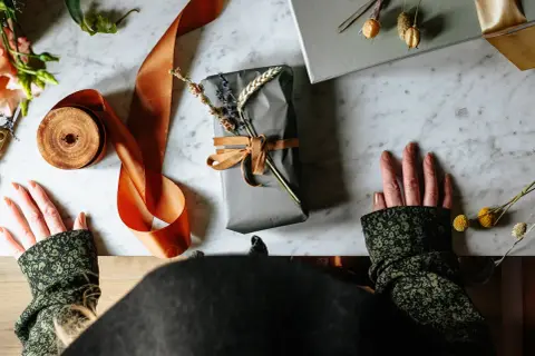 Händer som förbereder sig för att slå in runda paket med grått papper, orange sidenband och torkade blommor.