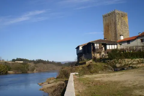 La torre de Lapela se alza junto a un río, con casas antiguas a su lado y un cielo azul despejado.