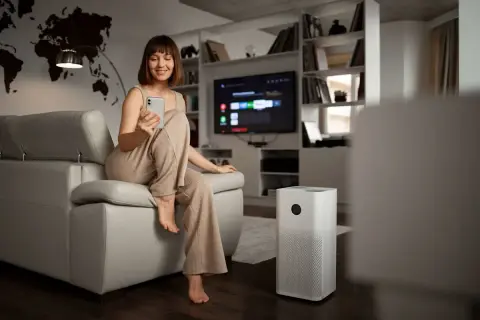 Donna sorride usando il telefono, accanto a un purificatore d'aria che garantisce la funzione deumidificatore.