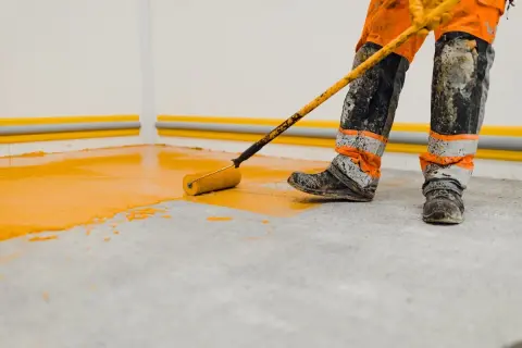 Un ouvrier en combinaison orange et noire utilise un rouleau pour peindre le sol d'un garage en jaune vif.