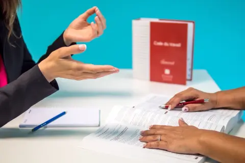 Dwie kobiety rozmawiają przy biurku, jedna wskazuje na otwartą książkę.
