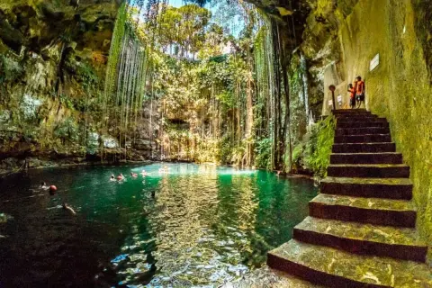Vista interior del cenote Chichén Itzá, con escaleras de piedra y nadadores en sus aguas turquesas, rodeado de vegetación colgante.