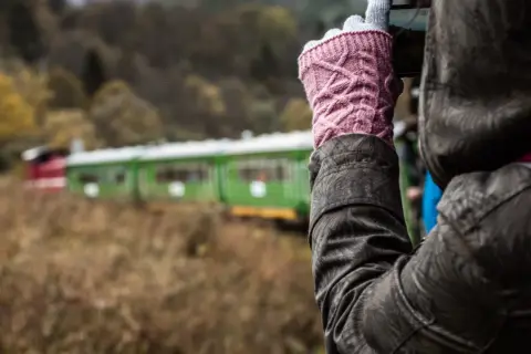 Osoba w różowych rękawiczkach fotografuje zielono-czerwony pociąg. Która kolejka bieszczadzka, która trasa ciekawsza?