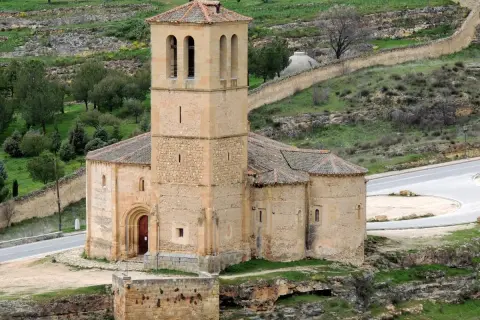 La iglesia de la Veracruz, de piedra y con un campanario robusto, se alza sobre un paisaje verde y ondulado, con un camino serpenteante a su lado.