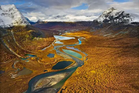 Herbstliche Farben im Tal mit Fluss und Seen, ideal für fernwanderwege schweden. Schneebedeckte Berge im Hintergrund.