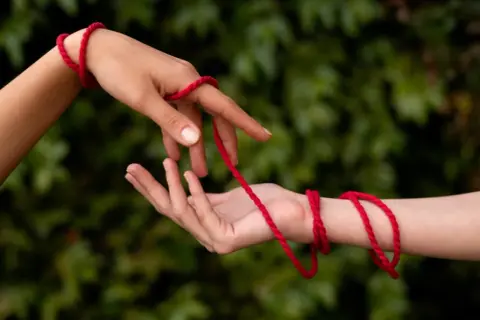 Dwie dłonie splatają czerwoną nitkę. Czy czerwona nitka to grzech? Symbol połączenia, losu, a może czegoś więcej?