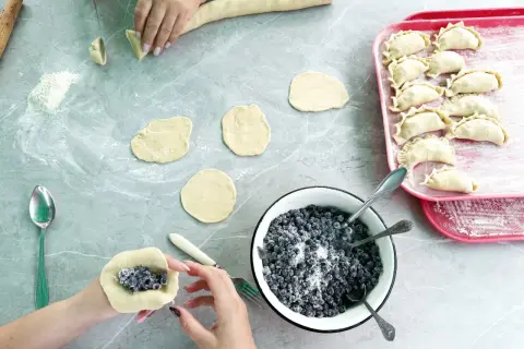 Ręce formują ciasto wonton z jagodami i cukrem. Na blacie leżą okrągłe placki ciasta, miska z jagodami i tacka z gotowymi pierożkami.