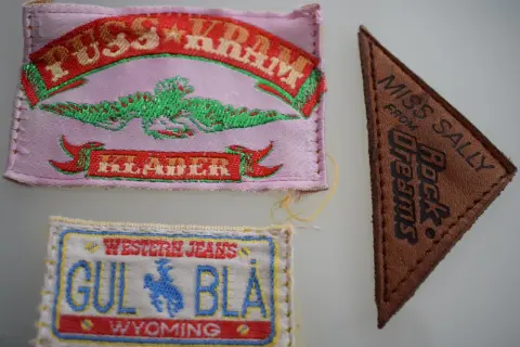 Tre märken: ett rosa med texten "PUSS & KRAM JEANS" och en grön drake, ett brunt triangelformat med "MISS SALLY FROM Rock Dreams", och ett vitt med "WESTERN JEANS GUL BLÅ WYOMING".