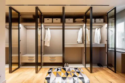 Armario empotrado moderno con puertas de cristal, perchas con ropa blanca y cajones.