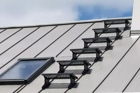 Sześć czarnych stopni bezpieczeństwa na metalowym dachu, prowadzących do okna dachowego. Solidne opierzenie dachu zapewnia bezpieczne przejście.