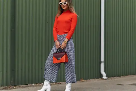 Kobieta w pomarańczowym golfie i kraciastych kulotach prezentuje stylowe kuloty stylizacje. Białe kozaki i torebka dodają charakteru.
