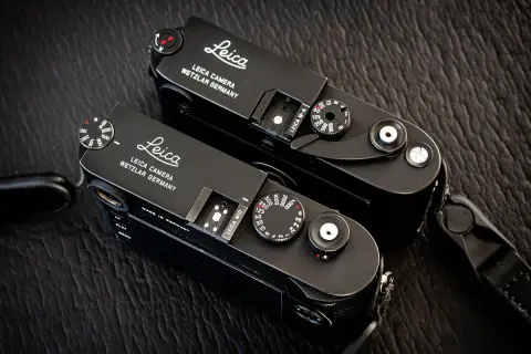 Two black Leica M cameras, a Leica M10 and a Leica M-A, sit side-by-side on a textured dark surface.