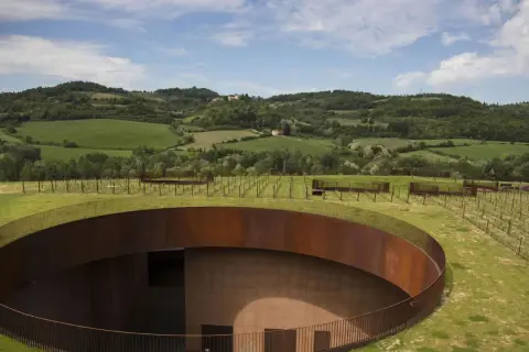 Un cerchio di metallo arrugginito emerge dal verde delle colline del Chianti, con vigneti che si estendono all'orizzonte sotto un cielo azzurro.