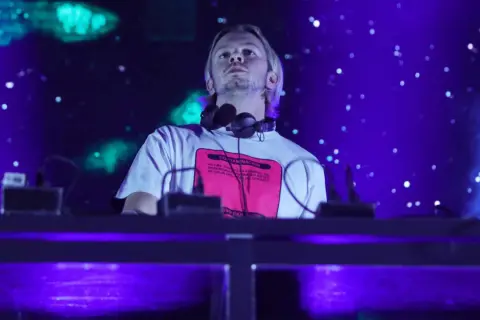 DJ na scenie, w słuchawkach na szyi, z napisem na koszulce. W tle niebieskie światła i efekty. To część lineupu Wisłoujście.