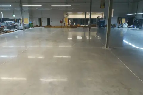Amplio espacio industrial con suelo de **cemento pulido** impecable, reflejando las luces del techo. Se aprecian montones de tierra y maquinaria en el fondo.