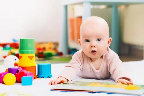Niemowlę leży na brzuchu obok kolorowych zabawek sensorycznych i otwartej książeczki.