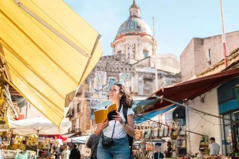 Turista sonriente con libro y cámara explora los vibrantes mercados de Palermo, con un domo histórico al fondo.
