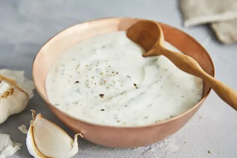 Ein cremiger Knoblauch Joghurt Dip in einer Kupferschale, garniert mit Pfeffer und Kräutern. Daneben liegen frische Knoblauchzehen.