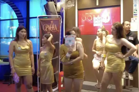 Kobiety w złotych sukienkach pozują przed banerem "Pacha". W tle neonowy napis "Shots". To scena z nocnego życia, być może związana z ping pong show w Tajlandii.