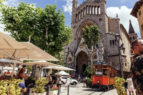 En charmig spårvagn passerar framför en kyrka i Soller, Palma. Folk sitter vid uteserveringar under parasoll.