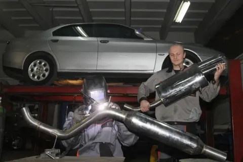 Mechanik w masce spawalniczej wykonuje spawanie wydechu. Obok stoi kolega z gotową częścią.