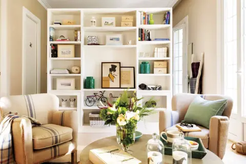 Acogedor salón con estanterías blancas llenas de libros y objetos decorativos, dos sillones de rayas y una mesa de centro con flores. Un rincón que evoca la calidez de las casas pequeñas.
