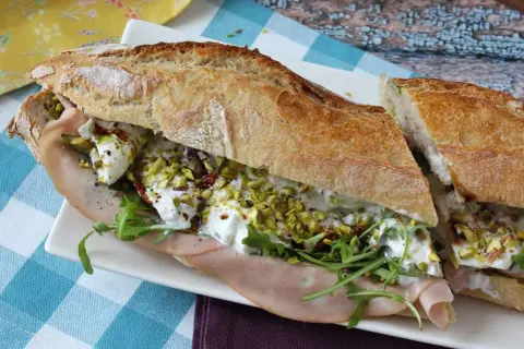 Un sandwich baguette garni de jambon, roquette, tomates séchées, pistaches concassées et burrata crémeuse.