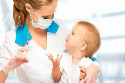 Pielęgniarka z maską i rękawiczkami trzyma strzykawkę z różowym płynem, przygotowując się do podania szczepionki 5w1 czy 6w1 dziecku.
