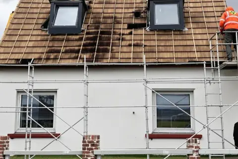 Gerüst am Haus, Dach wird saniert. Ein Arbeiter in Warnweste prüft die Arbeiten. Wer kontrolliert Sanierungspflicht?