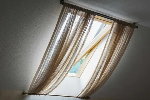 Voilage léger sur un montage store Velux ancien modèle, laissant passer la lumière du jour.