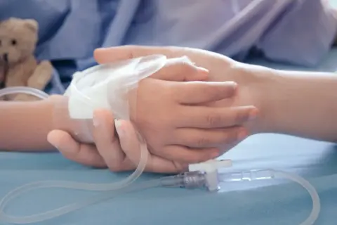 Dłoń dorosłego obejmuje dłoń dziecka z wenflonem. W tle pluszowy miś. Dokumentacja do orzeczenia o niepełnosprawności i zasiłek pielęgnacyjny.