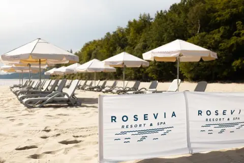 Plaża z leżakami i parasolami, w tle las, na pierwszym planie biały baner z logo "ROSEVIA RESORT & SPA".