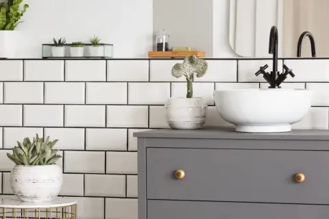 Planifier la dimension vasque salle de bain : un meuble gris, une vasque blanche, un robinet noir et des plantes vertes sur fond de carrelage blanc.