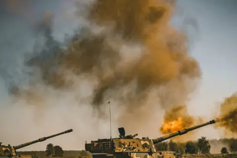 Dwa czołgi strzelają, tworząc kłęby dymu i ognia. Scena wygląda jak ćwiczenia, ale można by pomyśleć o "friendly fire".
