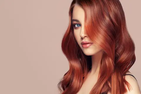 Ein Porträt einer Frau mit voluminösen, dunkelroten Haaren, die ihr Gesicht umspielen. Ihre blauen Augen blicken nachdenklich in die Ferne.