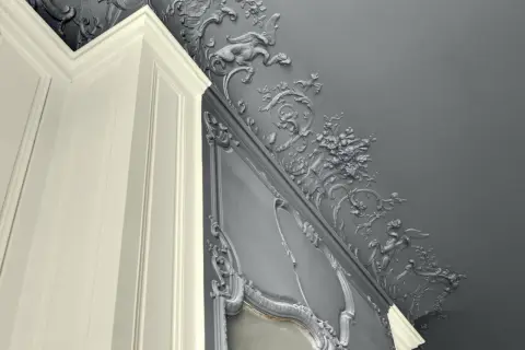 Magnifique peinture velours plafond avis : moulures grises ornées de créatures mythiques et de fleurs, contrastant avec un coin de mur blanc.