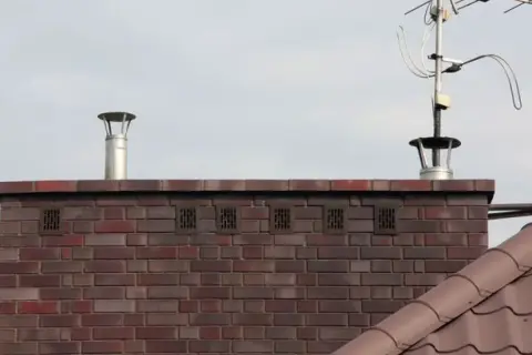 Zakończenie komina wentylacyjnego z metalową nasadką i kratkami wentylacyjnymi w ceglanej ścianie. Obok widoczna antena telewizyjna.