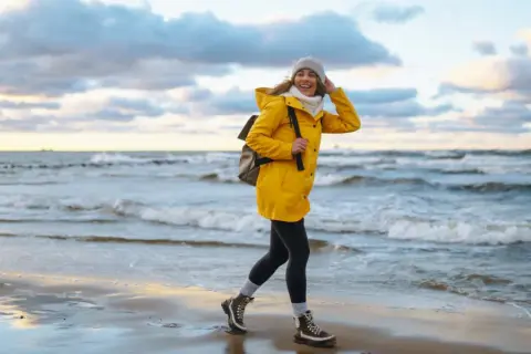 Uśmiechnięta kobieta w żółtej kurtce spaceruje po plaży. Wiatr rozwiewa jej włosy. To piękny jesienny dzień nad morzem.