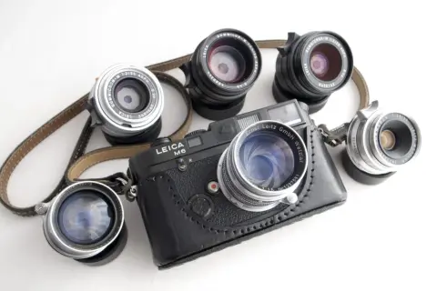 A Leica M6 camera with a leather case is surrounded by several best M mount lenses, showcasing a collection of classic photographic gear.