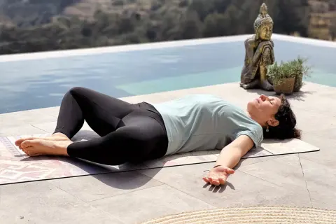 Mujer en posición de savasana, disfrutando de los beneficios de la relajación junto a una piscina, con una estatua de Buda de fondo.