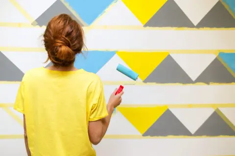 Frau mit rotem Dutt streicht mit Roller geometrische Muster an Wand. Ideen für Wand streichen mit Muster abkleben.