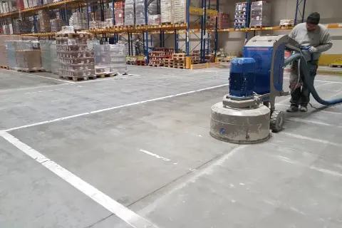 Pracownik wykonuje szlifowanie betonu w magazynie, używając maszyny z odkurzaczem.