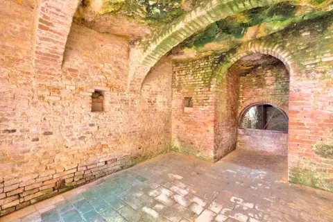 Interno di Santa Maria a Monte: muri in mattoni grezzi, volte muschiose e un arco con una finestra semicircolare.
