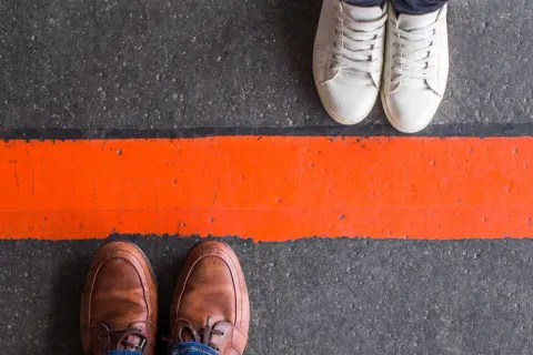 Dwie pary butów, jedna elegancka, druga sportowa, stoją po przeciwnych stronach pomarańczowej linii. Jak radzić sobie z zawiścią innych?