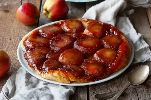 En gyllene tarte tatin med karamelliserade äpplen på en vit tallrik, omgiven av äpplen och en linneduk.
