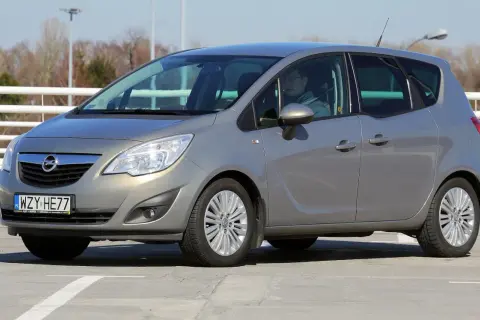 Szary Opel Meriva z kierowcą za kierownicą, zaparkowany na betonowym placu.