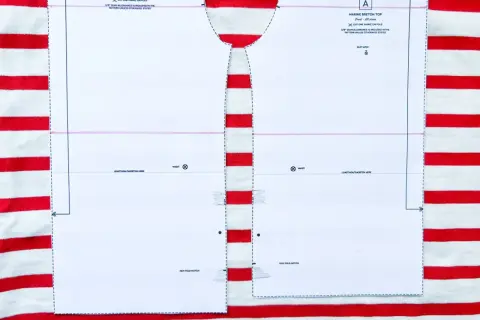 Wzór wykroju na bluzkę typu bretońskiego leży na materiale w biało-czerwone paski. Dzianina w paski idealnie pasuje do morskiego stylu.