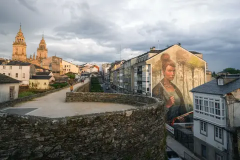 Mural de una mujer en la muralla de la fortaleza Lugo, con la catedral al fondo y un cielo nublado.