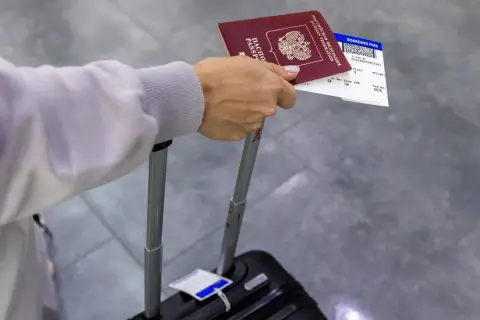 Dłoń trzyma paszport i kartę pokładową, która jest biletem na samolot. Obok walizka.