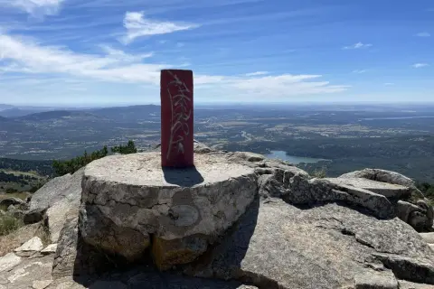 Pilar rojo con grafitis en la cima de una montaña, con un paisaje de valle y lago al fondo. La **cabeza lijar** del pilar parece desgastada por el tiempo.